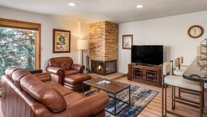 A living room with a stone fireplace, leather sofa, and a TV. - Summer Haven in Snowmass! Stunning Balcony Views, Pool/Hot Tub, Wood FP, Sleeping Den, W/D & Parking (Snowmass Village)