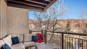 A balcony with a sofa and a table, overlooking a snowy landscape with trees and hills. - Summer Haven in Snowmass! Stunning Balcony Views, Pool/Hot Tub, Wood FP, Sleeping Den, W/D & Parking (Snowmass Village)