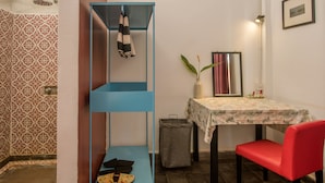 Um banheiro com azulejos estampados, uma prateleira azul, uma cadeira vermelha, uma mesa com toalha de mesa florida e um quadro na parede.