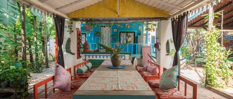 Un espace extérieur abrité, avec une table et des chaises, entouré de plantes et d’un bâtiment aux couleurs vives en arrière-plan.