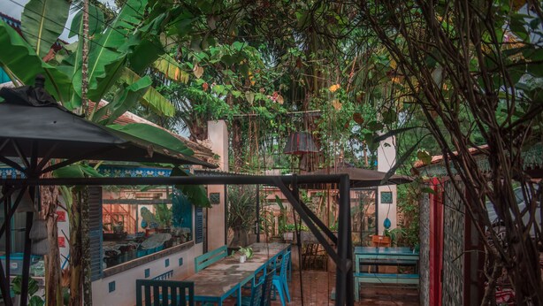 Um pátio externo com uma mesa comprida e cadeiras, rodeado por vegetação exuberante e um chão de azulejos.