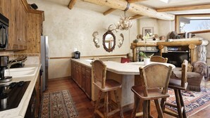 A rustic kitchen with wooden beams, a central island with stools, a fireplace, and a stone wall. - Beautiful Snowmass Getaway! Scenic Views, Balcony, Pool, Hot Tub, Gas FP, Elevator & Trail Access (Snowmass Village)