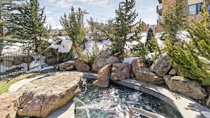 A hot tub surrounded by rocks and evergreen trees, with a snow-covered ground and a building in the background. - Beautiful Snowmass Getaway! Scenic Views, Balcony, Pool, Hot Tub, Gas FP, Elevator & Trail Access (Snowmass Village)