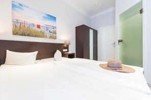 Una cama bien hecha con sábanas blancas, cabecera de madera, mesita de noche con lámpara, armario y una puerta con marco verde.