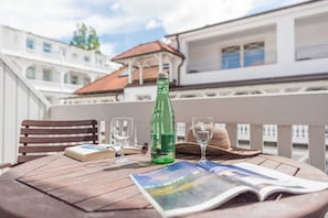 Ett träbord med en flaska, glas, en bok och en hatt på en balkong med vita räcken och byggnader i bakgrunden.