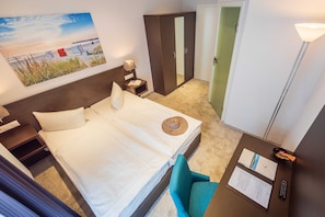 A hotel room with a bed, a desk with a lamp, a chair, a wardrobe, and a painting on the wall. - Double room with balcony - Hotel Villa Belvedere (Binz)