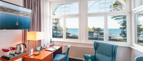 Een hotelkamer met een groot raam, een bureau met lamp, een televisie en twee blauwe fauteuils.