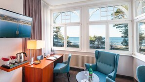 Una habitación de hotel con un ventanal grande, un escritorio con lámpara, un televisor y dos sillones azules.