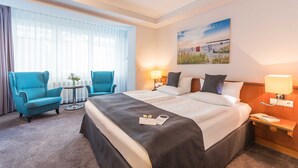 Quarto de hotel com uma cama grande, duas mesas de cabeceira, uma escrivaninha, uma cadeira e uma janela com cortinas translúcidas.