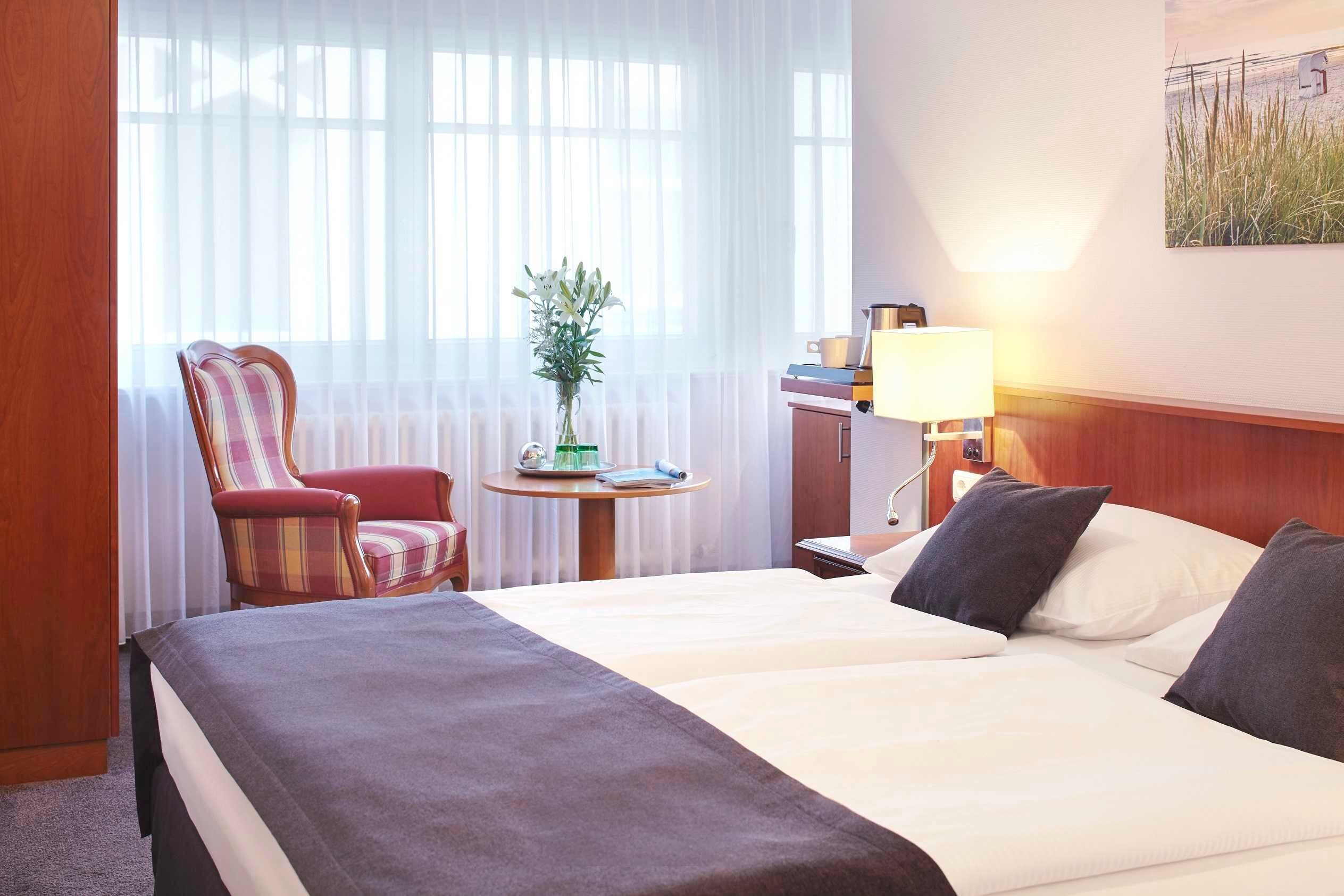 Una habitación de hotel con una cama, una silla, una mesita con una planta, una lámpara y un cuadro en la pared.