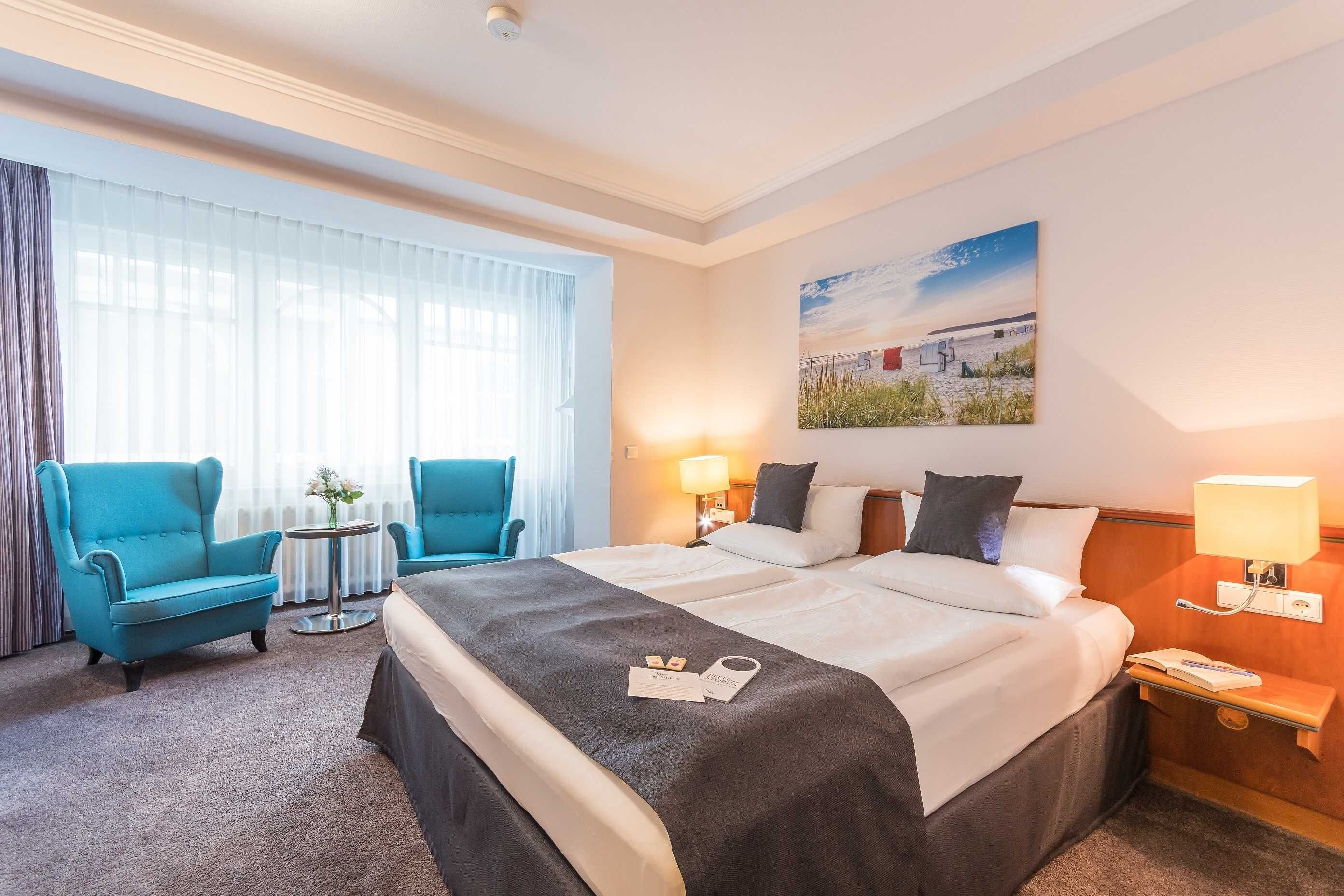 A hotel room with a large bed, two bedside tables, a desk, a chair, and a window with sheer curtains.