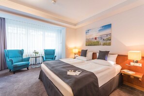 A hotel room with a large bed, two bedside tables, a desk, a chair, and a window with sheer curtains.