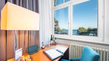 Habitación de hotel con escritorio, una laptop, un control remoto, una silla, una lámpara, ventana con vista al mar y una mesita con una botella y vasos.