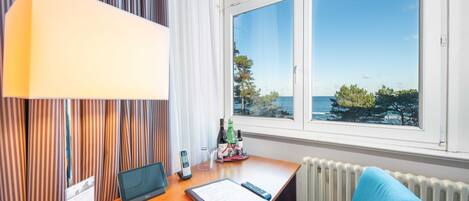 A hotel room with a desk, a laptop, a remote control, a chair, a lamp, a window with a view of the sea, and a small table with a bottle and glasses.