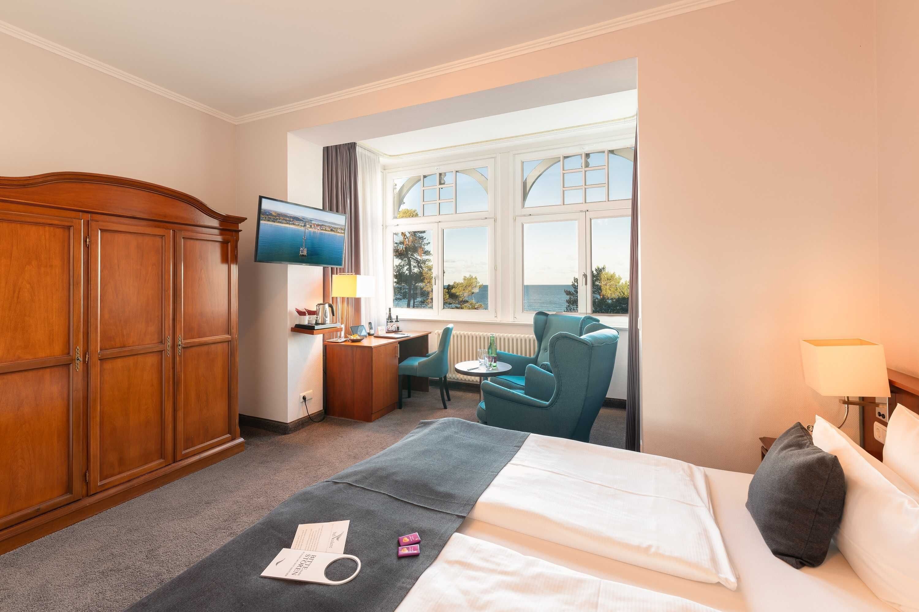 A hotel room with a bed, a desk, a chair, a TV, and a window with a view of the sea.