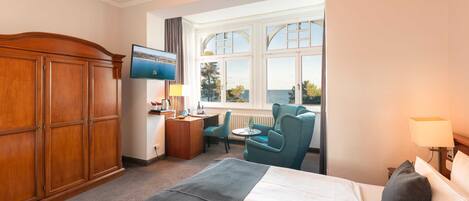 A hotel room with a bed, a desk, a chair, a TV, and a window with a view of the sea.