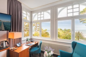 A hotel room with a desk, two chairs, a TV, a window with a view of trees and the sea, and a radiator. - Junior suite with panoramic sea view - Hotel Villa Belvedere (Binz)