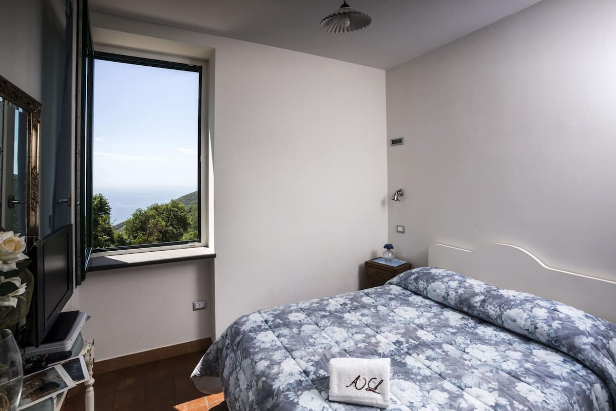 A bedroom with a large window, a bed with floral bedding, a bedside table with a lamp, and a view of greenery outside.