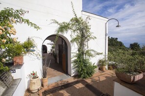 A white building with a covered patio area, featuring a metal door, potted plants, and a street lamp. - Villa Lupara (Salerno)