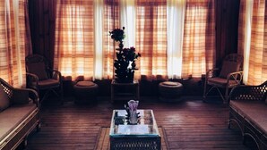 Een gezellige kamer met houten vloeren, een centrale salontafel, rieten meubels en een groot raam met gordijnen.