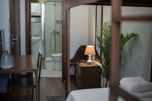 A hotel room with a wooden desk, a chair, a bedside table with a lamp, a bed, a potted plant, and a view of the bathroom. - Casa do Teatro (Ponta Delgada)