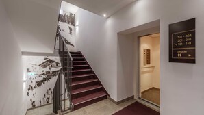 Un escalier avec une rampe en verre, un mur orné d’une fresque photographique en noir et blanc, et un panneau indiquant les numéros des chambres ainsi que le symbole des toilettes.