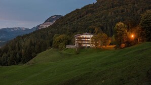 Un immeuble à plusieurs étages, doté d’un toit plat, entouré d’un paysage verdoyant et d’une colline boisée.