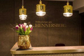 A reception desk with a gold vase of flowers, three gold pendant lights, and a wall with the text 'Willkommen am HENNERSBERG'.
