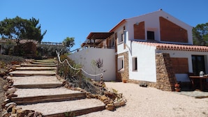 Une maison à deux étages, avec des fondations en pierre, un toit recouvert de tuiles rouges et une allée de gravier menant à un escalier en bois.