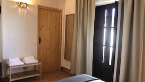Une chambre à coucher avec un lit, un banc en bois sur lequel sont posées des serviettes, une lampe fixée au mur et une porte avec des rideaux.