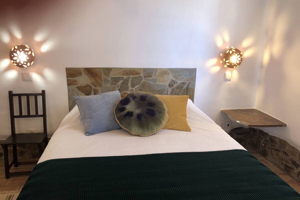 Ein Bett mit Stein-Kopfteil, zwei Wandleuchten, ein Sessel und ein kleines Holzregal.