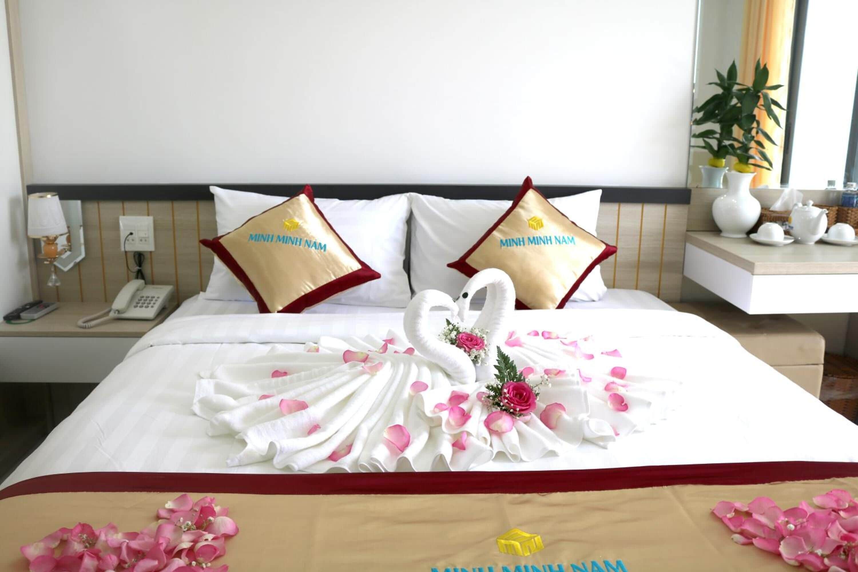 A hotel room with a neatly made bed, two pillows with the hotel's name, a swan-shaped towel arrangement, and a bedside table with a lamp and phone.