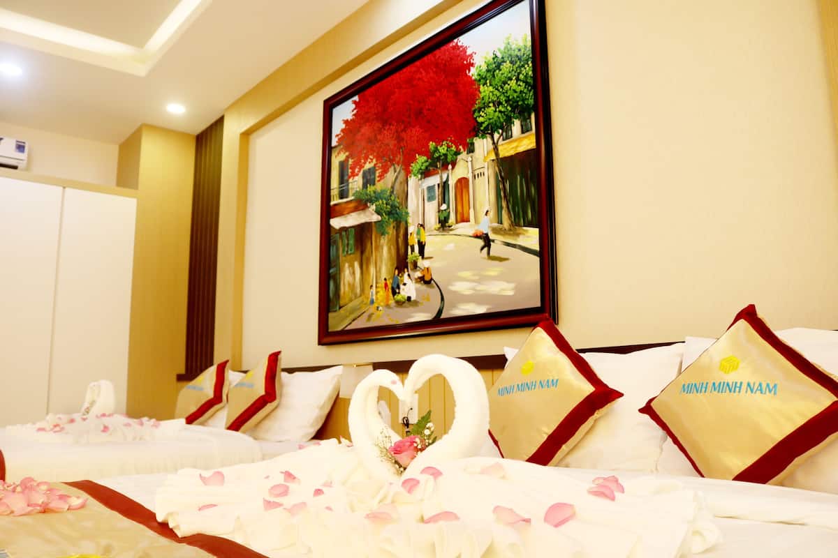 Quarto de hotel com uma cama decorada com pétalas de rosa e um adorno em formato de coração. Há uma pintura de uma cena urbana na parede.