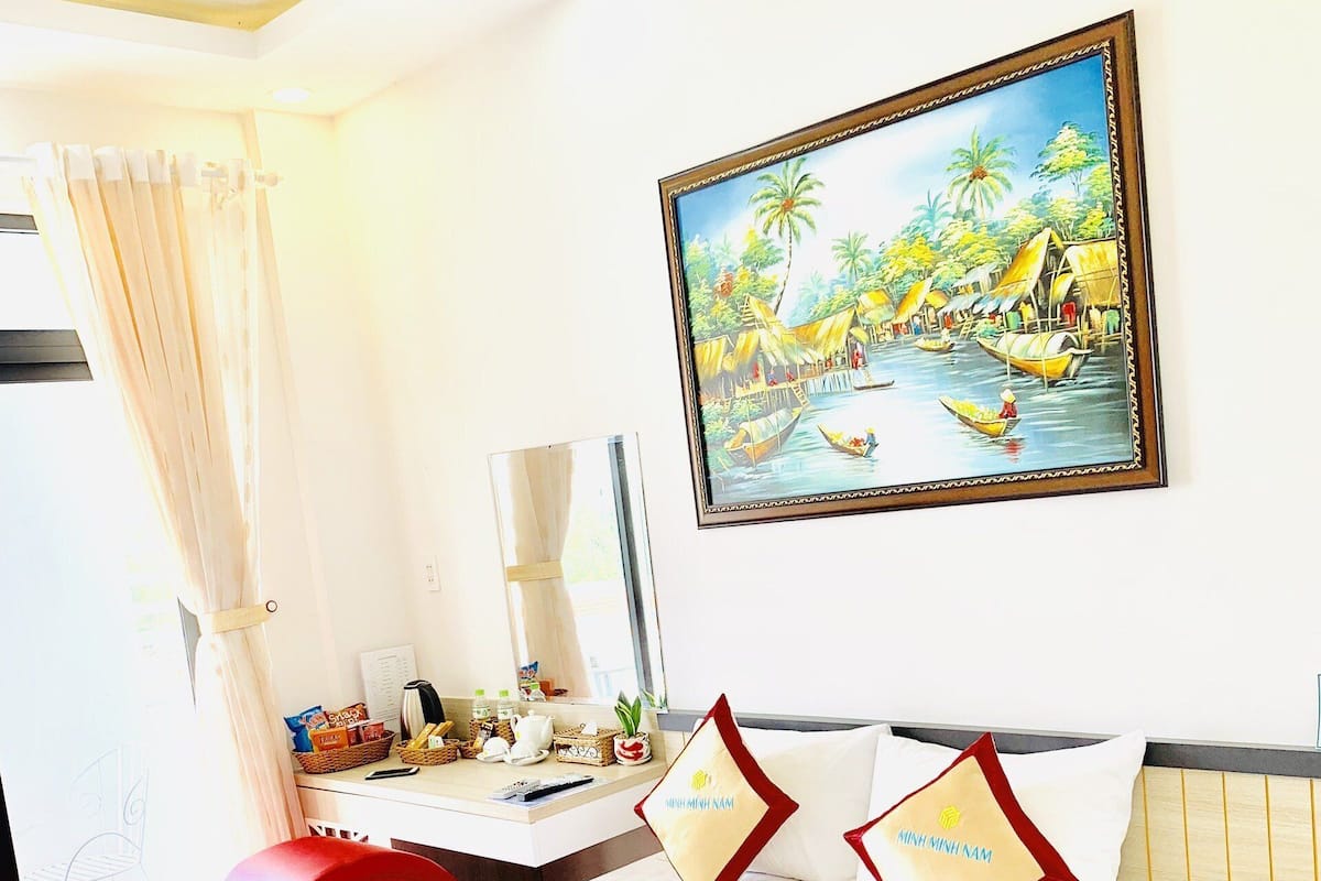 Quarto de hotel com cama, escrivaninha, cadeira, janela com cortinas, quadro de uma paisagem tropical e almofada com texto.