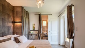 A bedroom with a large bed, a bedside table, a nightstand, a chandelier, a bathroom visible through an open door, and a window with curtains. - Albizia - Charming gîte in the heart of Perigord - Max 4 people (Nouvelle-Aquitaine)