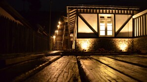 A wooden deck with a staircase leading to a building with a stone facade and a window with lights. - Loversleap Cottage (Nuwara Eliya)