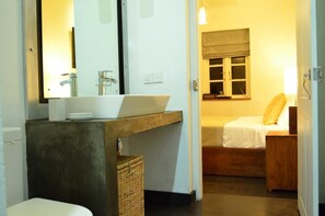A bathroom with a white sink, a mirror, and a wooden vanity. There is a bed with a white mattress and a brown headboard in the adjacent room.