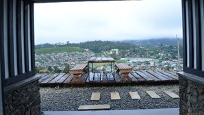 A view from a covered outdoor area with a wooden table and benches, overlooking a town with houses and greenery. - Loversleap Cottage (Nuwara Eliya)