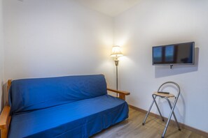 A small room with a blue sofa, a standing lamp, a TV on the wall, and a small folding chair. - GuestHost - Modern APT just 2 steps from the Ciclovia! x3 (Tarvisio)
