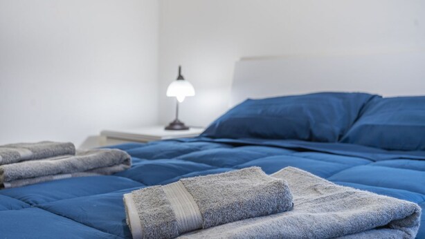 A neatly made bed with blue bedding, a lamp on the nightstand, and folded towels. - GuestHost - Modern APT just 2 steps from the Ciclovia! x3 (Tarvisio)
