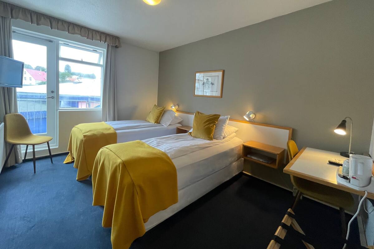 Ein Hotelzimmer mit zwei Betten, einem Schreibtisch, einem Stuhl und einem Fenster mit Blick auf Gebäude.