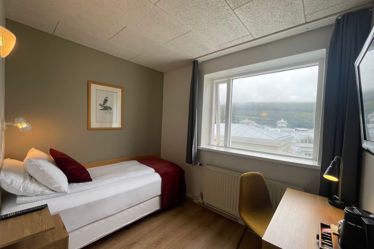 Ein Hotelzimmer mit Bett, Schreibtisch, Stuhl, Fenster mit Blick und Heizkörper.