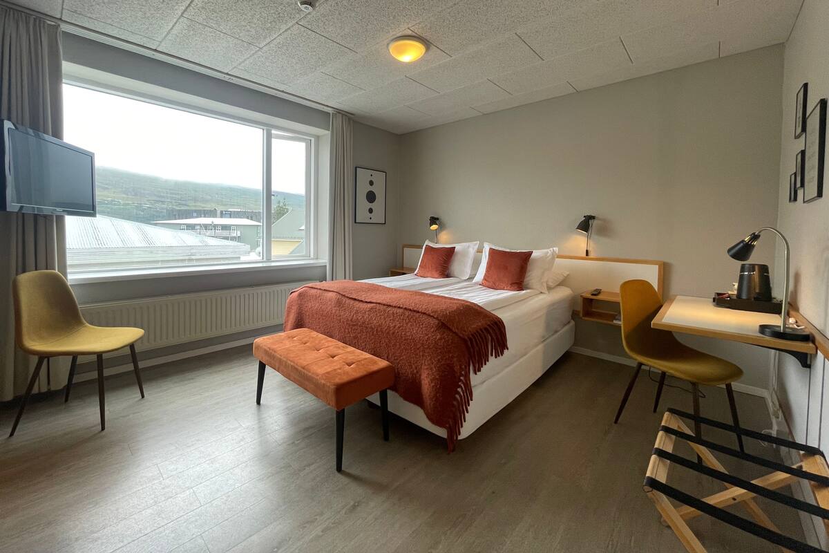 Ein Hotelzimmer mit Bett, zwei Stühlen, einem Schreibtisch, einem Fernseher und einem Fenster mit Blick auf Gebäude und Hügel.