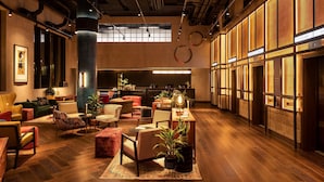 Un vestíbulo amplio con techo alto, suelo de madera y diversas opciones de asientos, incluyendo sofás y sillones. Varias plantas en macetas y un pilar grande en el centro.