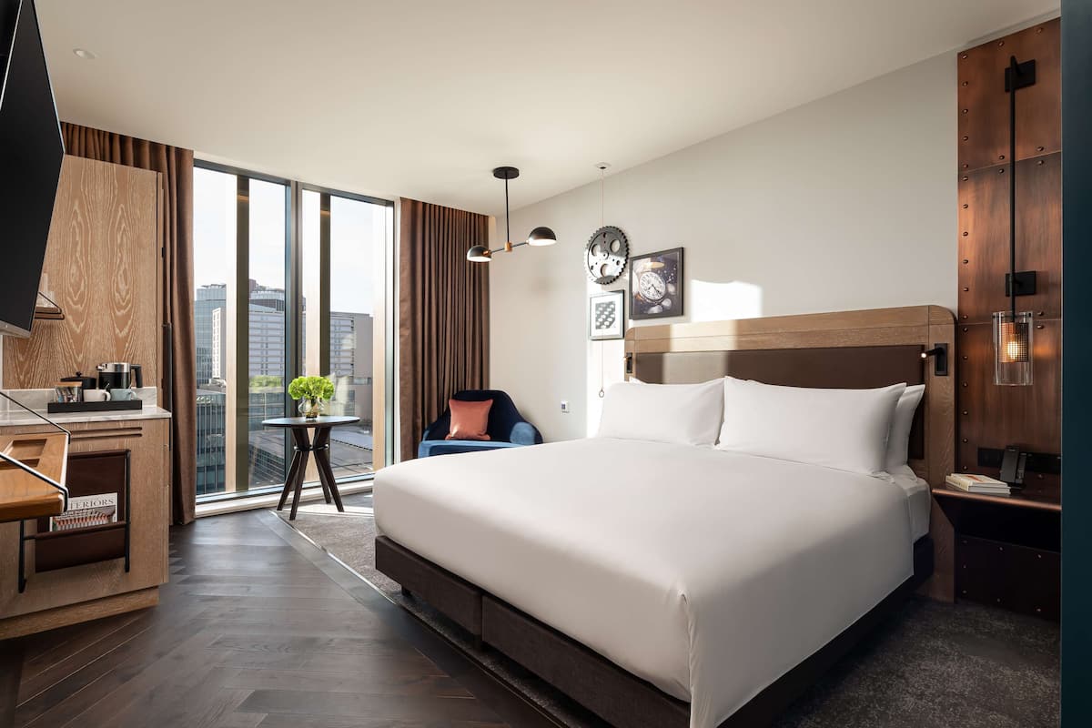 A modern hotel room with a large bed, a desk, a chair, and a view of the city through large windows.
