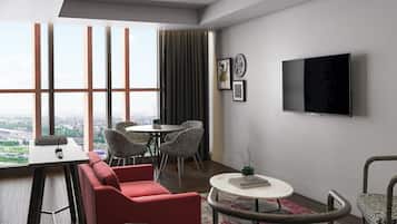 Una habitación de hotel moderna con un ventanal grande, un televisor de pantalla plana, una zona de comedor con mesa y sillas, y un sofá rojo.