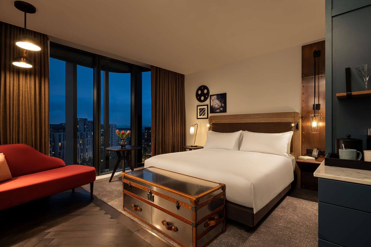 A modern hotel room with a large bed, a red sofa, a wooden chest of drawers, and a city view.