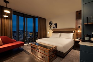 A modern hotel room with a large bed, a red sofa, a wooden chest of drawers, and a city view. - The Gantry London, Curio Collection by Hilton (London)