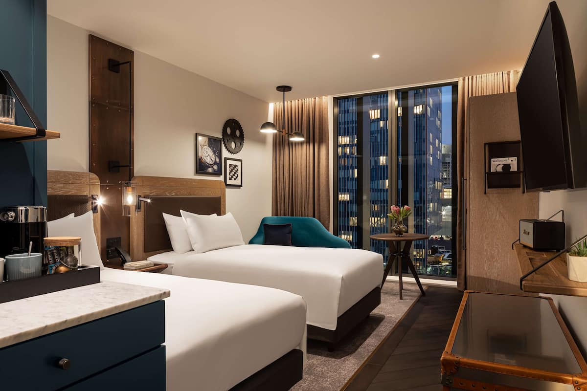 A hotel room with two beds, a small table, a TV, and a view of the city.