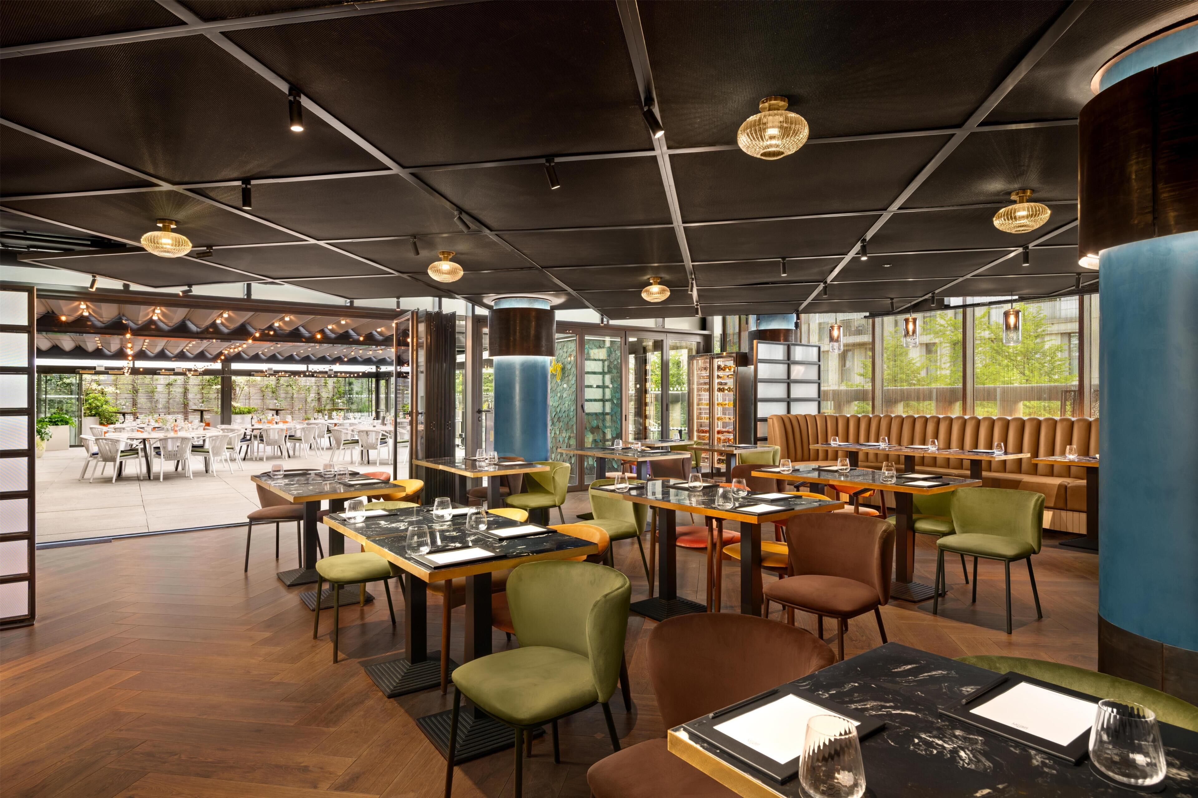 A restaurant with tables and chairs, large windows, and hanging lights.